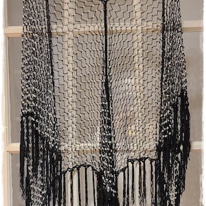 May include: Black, open-weave shawl with fringe. The shawl is made of a net-like material with a geometric pattern. It is hanging on a decorative gold-colored hanger.