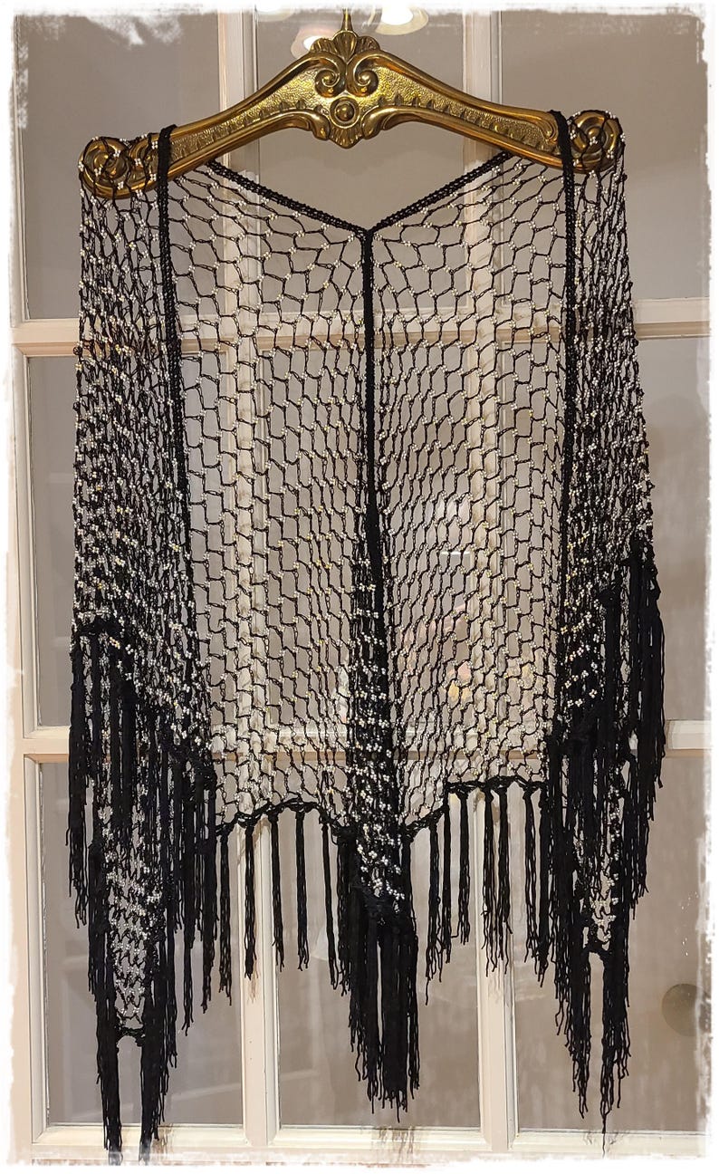 May include: Black, open-weave shawl with fringe. The shawl is made of a net-like material with a geometric pattern. It is hanging on a decorative gold-colored hanger.