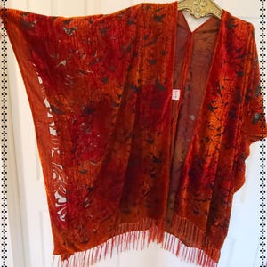 May include: A burnt orange velvet kimono with a floral pattern and fringe trim. The open-front garment is hanging on a gold hanger against a white door. The fabric has a textured appearance and a dark floral design.