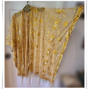 May include: A sheer gold kimono-style cover-up featuring an intricate pattern of embroidered stars, sunbursts, and crescent moons. The garment is embellished with gold sequins and beads throughout, with a scalloped edge along the hemline.