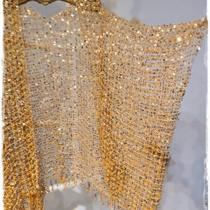 May include: A gold sequin shawl with a net-like design. The shawl is draped over a gold hanger, showcasing its shimmering texture and intricate pattern. The sequins catch the light, creating a dazzling effect.