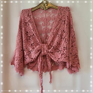 May include: A dusty rose-colored lace bolero featuring an intricate floral pattern, wide three-quarter length sleeves, and a front tie closure. The garment is constructed from a delicate, open-work crochet fabric with scalloped edges.