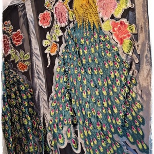 Puede incluir: Prenda negra y gris con un gran dise&ntilde;o de pavo real. El pavo real presenta una cabeza amarilla, plumas verde azulado con acentos rosas y amarillos, y est&aacute; rodeado de bordados florales rosas y rojos. La prenda tiene una textura aterciopelada.