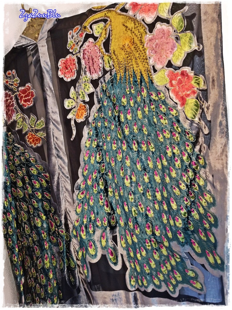Puede incluir: Prenda negra y gris con un gran dise&ntilde;o de pavo real. El pavo real presenta una cabeza amarilla, plumas verde azulado con acentos rosas y amarillos, y est&aacute; rodeado de bordados florales rosas y rojos. La prenda tiene una textura aterciopelada.