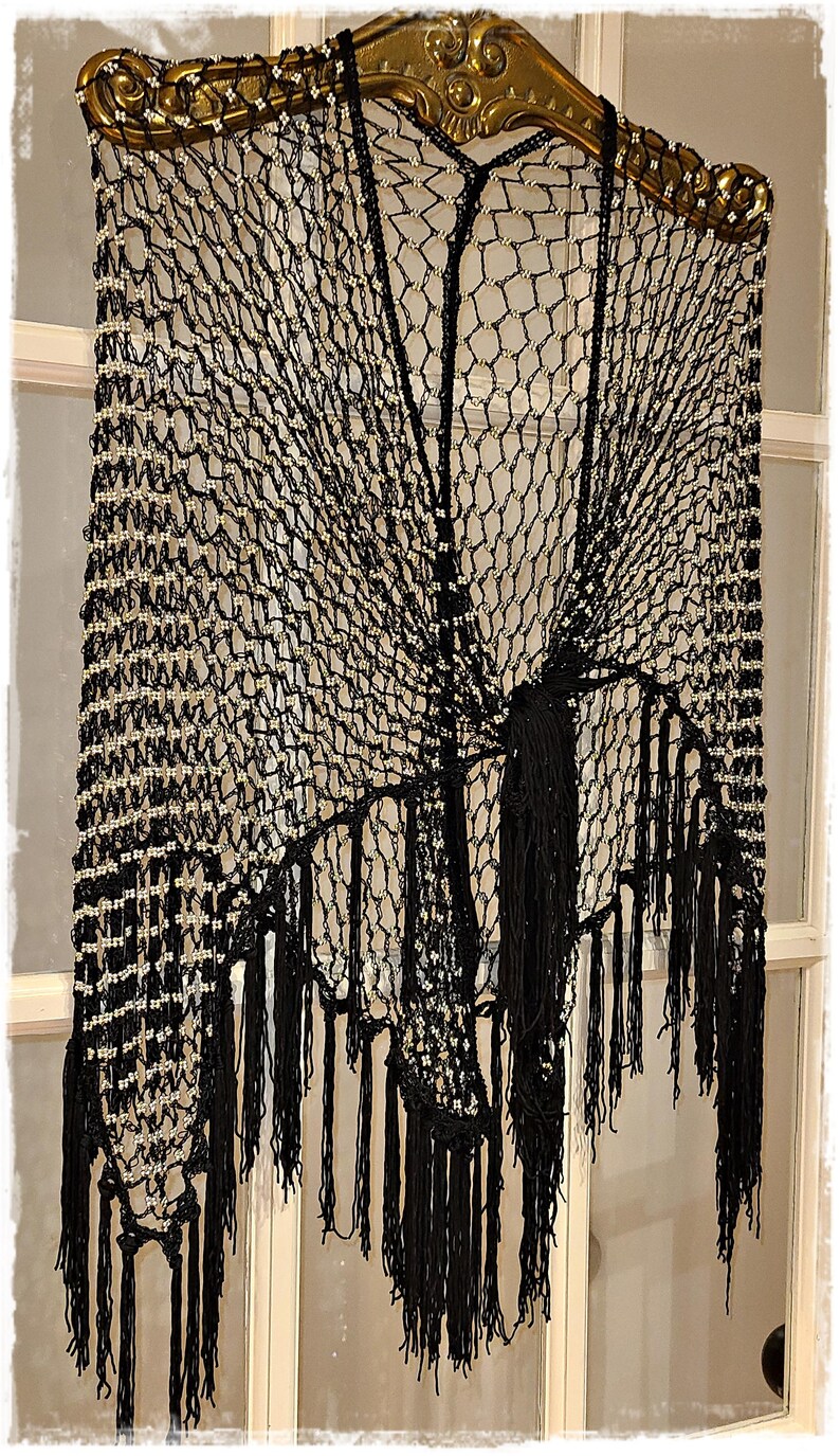 May include: Black crocheted shawl with fringe, adorned with small, light-colored beads. The shawl is displayed on a decorative gold-colored rod. The intricate net-like design and long fringe create a vintage, elegant look.