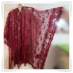 May include: A sheer, burgundy-colored kimono-style wrap with an intricate pattern of sequins and embroidered designs. The wrap features a celestial motif with stars, moons, and floral elements. The fabric is lightweight and appears to be semi-transparent.