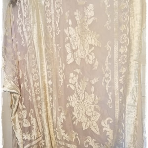 May include: A cream-colored sheer kimono featuring a velvet floral pattern and vertical decorative borders. The garment includes a fringe trim along the bottom edge and displays the text ZzaZuuBlu in the upper left corner of the image.
