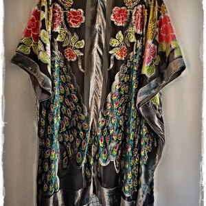 Puede incluir: Un kimono gris oscuro con ribete de terciopelo y flecos de cuentas. El kimono presenta un dise&ntilde;o floral y de plumas de pavo real en rojo, amarillo, verde y rosa. La prenda est&aacute; colgada sobre un fondo gris claro.