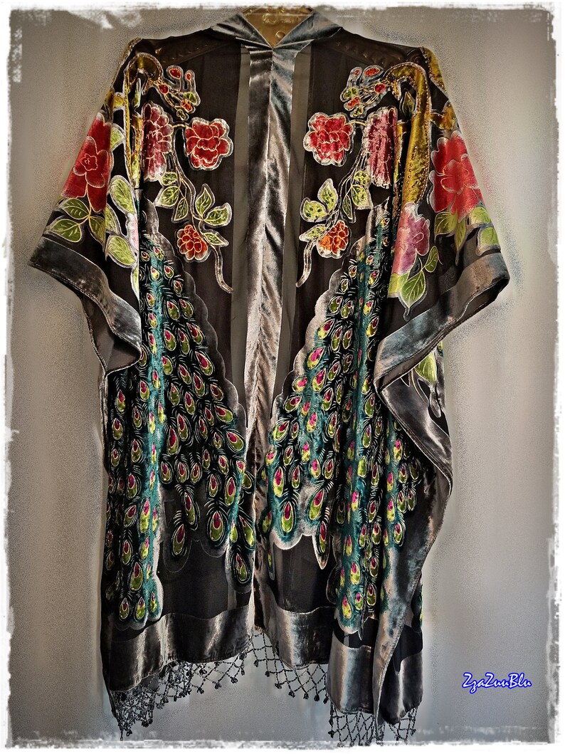 Puede incluir: Un kimono gris oscuro con ribete de terciopelo y flecos de cuentas. El kimono presenta un dise&ntilde;o floral y de plumas de pavo real en rojo, amarillo, verde y rosa. La prenda est&aacute; colgada sobre un fondo gris claro.