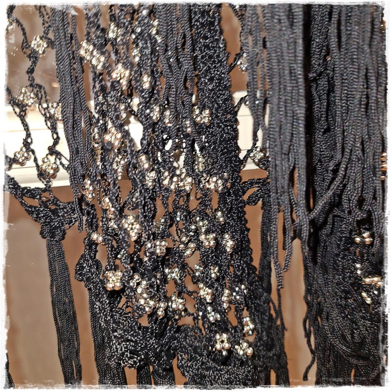 May include: Close-up of a black, fringed textile with intricate detailing. The fabric features a delicate, open-weave pattern adorned with clusters of small, silver-colored beads. The texture is a mix of smooth and fringed elements.