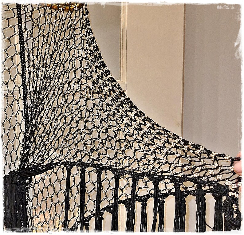May include: Black and silver crocheted shawl with a net-like design. The shawl features a fringe along the bottom edge and is embellished with silver accents. The top edge has a decorative beaded trim.