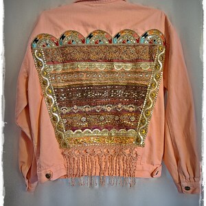 May include: A peach-colored denim jacket with elaborate embellishments. The back features a large, decorative panel with intricate beadwork, sequins, and fringe. The design incorporates gold, red, and brown tones, creating a bohemian aesthetic. The jacket is hanging on a hook.