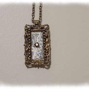 May include: A rectangular pendant with an ornate, antique gold-colored frame. The pendant features a white lace design set against a black background, with a small, round, copper-colored accent in the center. The pendant hangs from a matching chain.