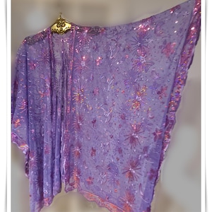 May include: A sheer, lavender-colored kimono-style garment with a floral and starburst pattern. The edges are trimmed with iridescent sequins. The garment is hanging on a gold-colored decorative hanger.