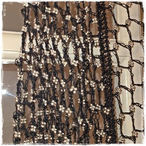 May include: Black open-weave scarf with silver-colored embellishments. The scarf has a delicate, lacy appearance, with small, sparkling accents evenly spaced throughout the fabric. The design suggests a formal or evening wear accessory.