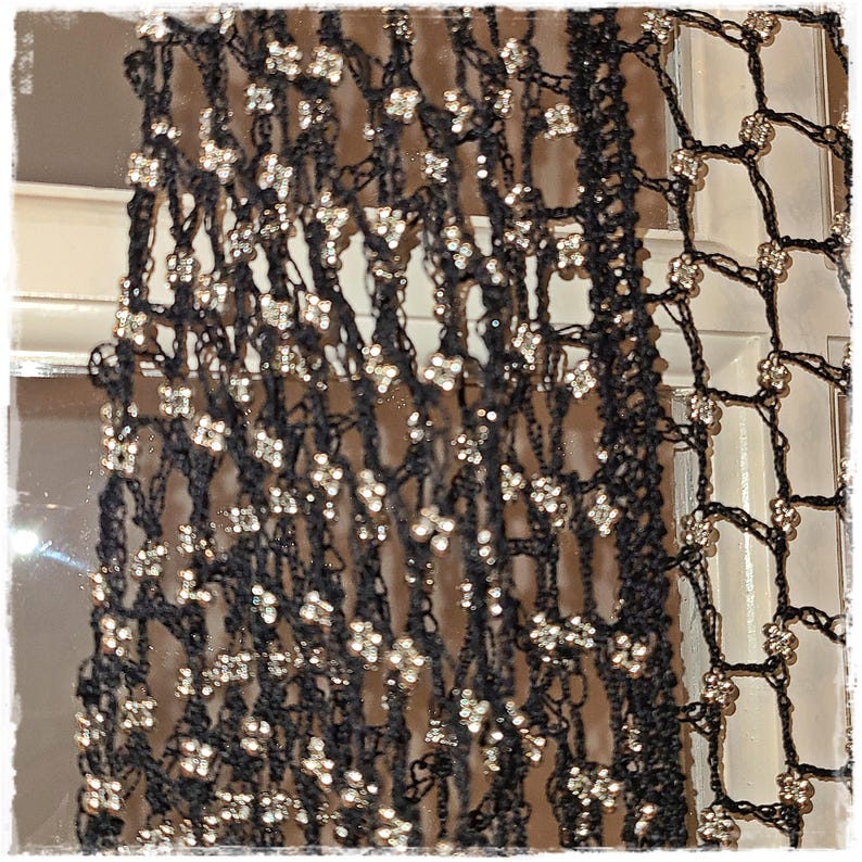 May include: Black open-weave scarf with silver-colored embellishments. The scarf has a delicate, lacy appearance, with small, sparkling accents evenly spaced throughout the fabric. The design suggests a formal or evening wear accessory.