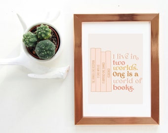 May include: A framed print with a copper-colored frame features the quote "I live in two worlds. One is a world of books." The print has a light peach background with a stack of book illustrations. A small cactus in a white ceramic holder is to the left.