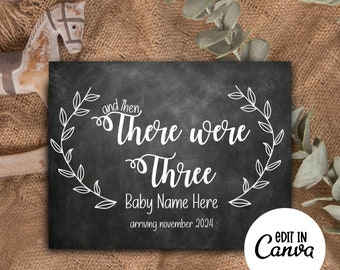 May include: Black chalkboard sign with white text announcing a new baby. The text reads "and then there were three Baby Name Here arriving November 2024". The sign is decorated with white leafy wreaths.