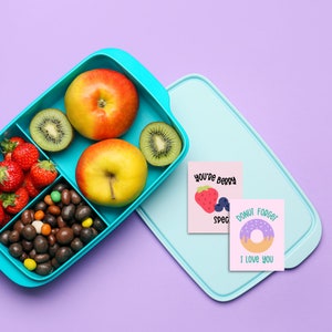 Op de afbeelding: Een blauwe lunchbox gevuld met verschillende snacks, waaronder fruit, snoep en kiwi. Twee kaartjes liggen bovenop de lunchbox met de tekst "You're Berry Special" en "Donut Forget I Love You".