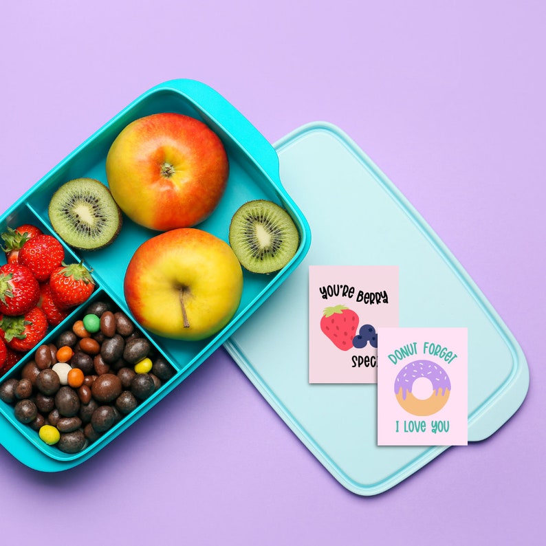 Op de afbeelding: Een blauwe lunchbox gevuld met verschillende snacks, waaronder fruit, snoep en kiwi. Twee kaartjes liggen bovenop de lunchbox met de tekst "You're Berry Special" en "Donut Forget I Love You".