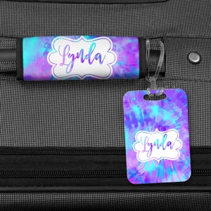 May include: A luggage tag and handle wrap with a tie-dye pattern in shades of blue, purple, and pink. The tag and wrap both have the name "Lynda" written in white cursive lettering inside a white frame.