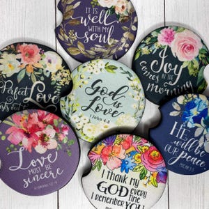 May include: A collection of car coasters with floral designs and inspirational quotes. The coasters feature various colors, including purple, blue, and white, with text such as "God is Love" and "It is well with my soul."