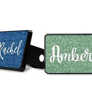May include: Two personalised hitch covers with glitter backgrounds. The left cover is blue with the name "Rachel" in white. The right cover is green with the name "Amber" in white.