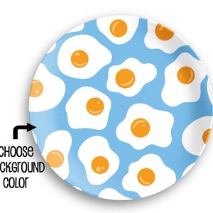 May include: A round plate with a light blue background featuring a repeating pattern of fried eggs. Each egg has a white egg white and a bright yellow yolk. Text on the left side says "CHOOSE BACKGROUND COLOR".