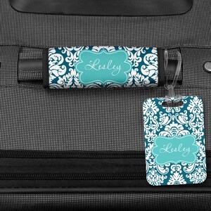 May include: A teal and white luggage tag with a damask pattern and the name "Lesley" in a teal frame. The tag is attached to a black suitcase with a teal and white luggage strap.