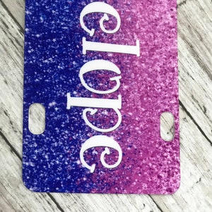K&ouml;nnte beinhalten: Ein lila und blauer Glitzerhintergrund mit dem Wort "Chloe" in wei&szlig;en Kursivbuchstaben. Der Hintergrund hat einen Verlaufseffekt, der von Lila zu Blau &uuml;bergeht. An den Seiten des Hintergrunds befinden sich zwei L&ouml;cher.