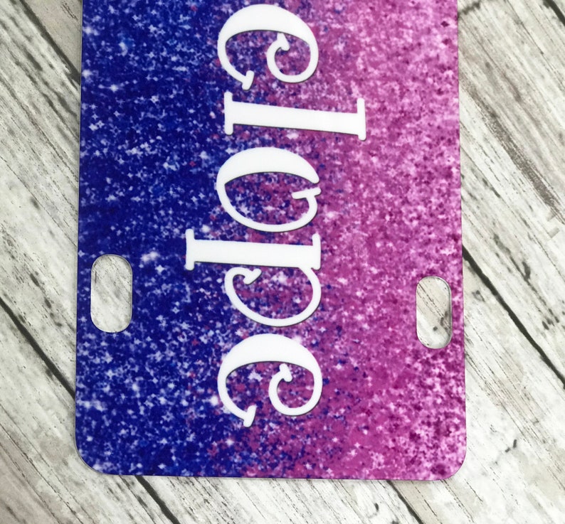 K&ouml;nnte beinhalten: Ein lila und blauer Glitzerhintergrund mit dem Wort "Chloe" in wei&szlig;en Kursivbuchstaben. Der Hintergrund hat einen Verlaufseffekt, der von Lila zu Blau &uuml;bergeht. An den Seiten des Hintergrunds befinden sich zwei L&ouml;cher.