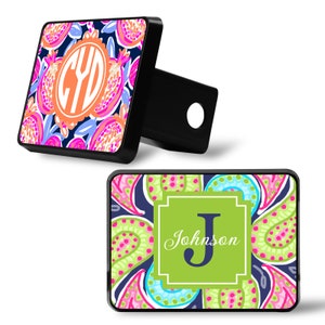 May include: Two black metal hitch covers with colorful patterned designs. The first cover features a monogram "CYD" in an orange circle on a navy blue background with pink, orange, and blue floral designs. The second cover features a green background with a white letter "J" and the text "Johnson" in a white cursive font. The background features a pink, green, and blue paisley design.