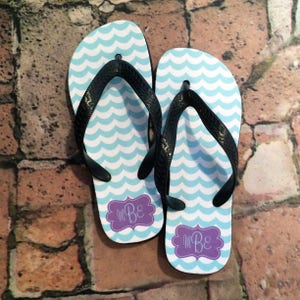 May include: A pair of blue and white flip flops with a wave pattern. The flip flops have a purple monogram that says "MB" on each strap.