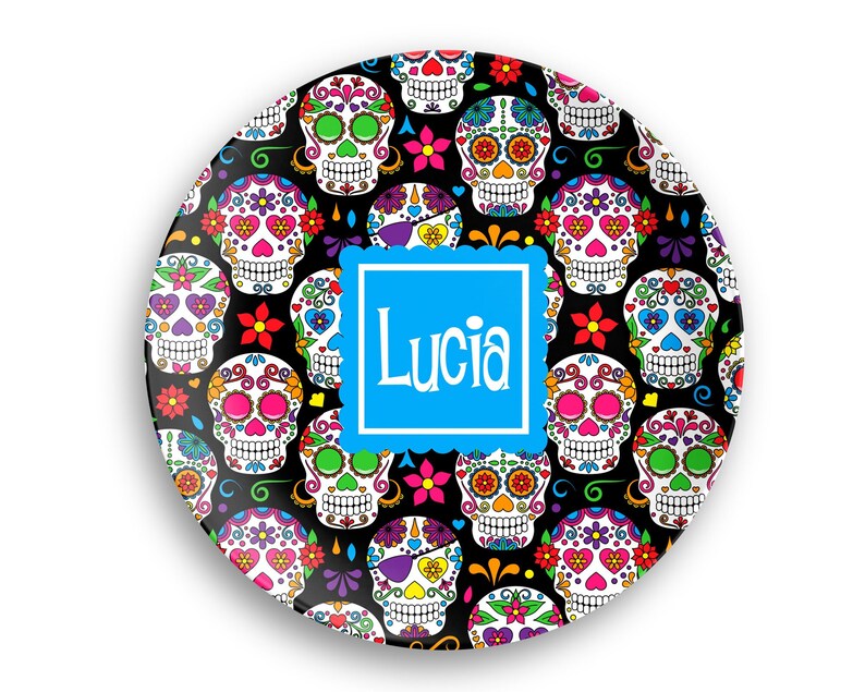 May include: A round decorative plate featuring a repeating pattern of colorful sugar skulls on a black background. A blue square in the center displays the name "Lucia" in white script. The skulls have various floral and heart designs.