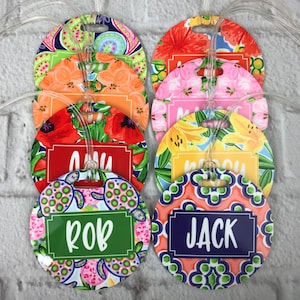May include: Eight colorful round luggage tags with different floral and geometric patterns. Each tag has a white rectangle with a name printed in black: Amy, Rob, Jack,  and others. The tags are attached to clear luggage straps.