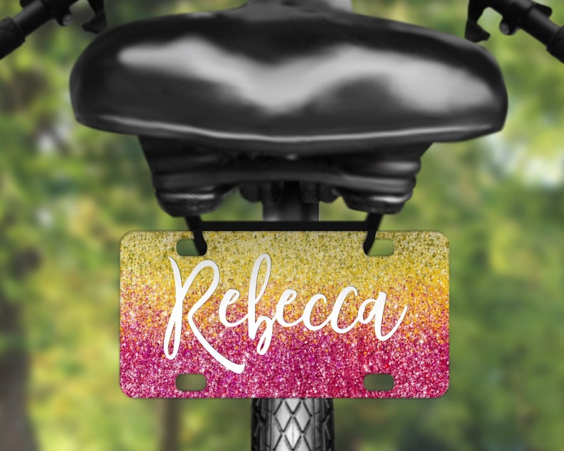 May include: A pink and yellow glitter bike licence plate with the name "Rebecca" written in white cursive font.