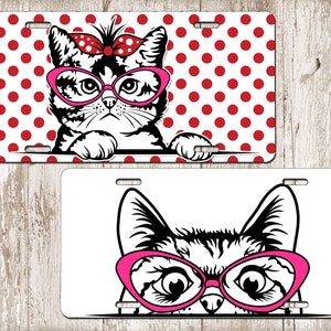 May include: Two license plates featuring cat designs. One plate has a black and white cat wearing pink glasses and a red bow, set against a red polka dot background. The other plate shows a cat's face with pink glasses.