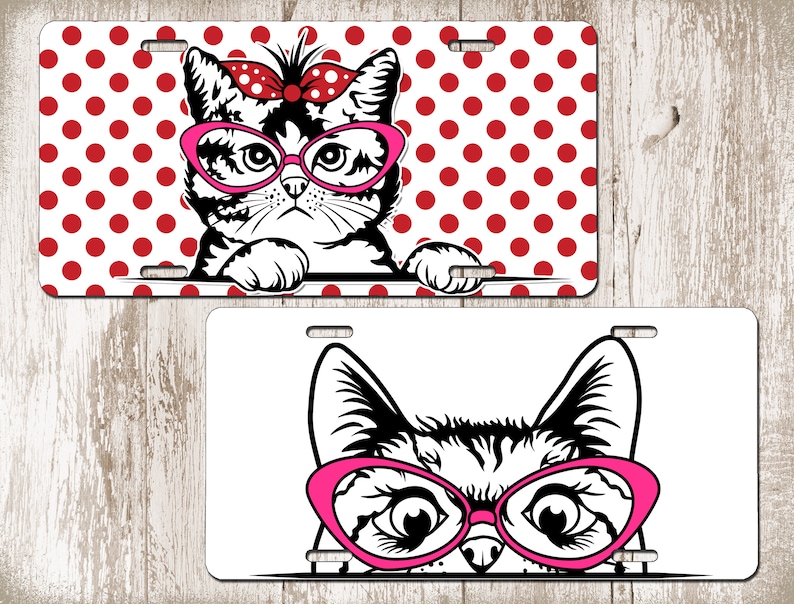 May include: Two license plates featuring cat designs. One plate has a black and white cat wearing pink glasses and a red bow, set against a red polka dot background. The other plate shows a cat's face with pink glasses.