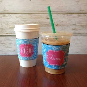 May include: Two coffee cups with personalized blue and pink patterned sleeves. The sleeve on the left cup has the monogram "RCH" and the sleeve on the right cup has the name "Lucy" in white lettering.