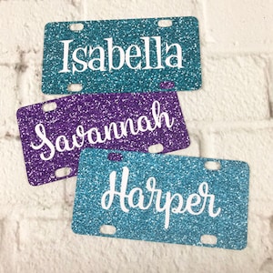 May include: Three glitter license plates with the names Isabella, Savannah, and Harper in white lettering.