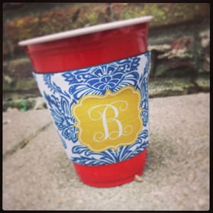 May include: A red plastic cup with a blue and white patterned fabric cup holder. The cup holder has a gold monogram with the letter "B" in a script font.