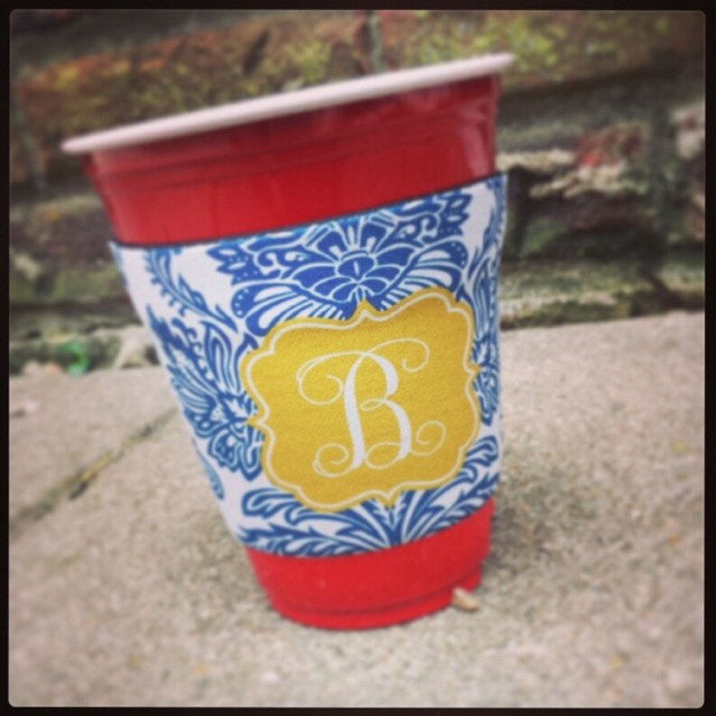 May include: A red plastic cup with a blue and white patterned fabric cup holder. The cup holder has a gold monogram with the letter "B" in a script font.