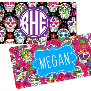 May include: Two colorful personalized license plates with a sugar skull design. The top plate has a black background with a purple monogram "BHE" in the center. The bottom plate has a pink background with the name "MEGAN" in a blue banner.