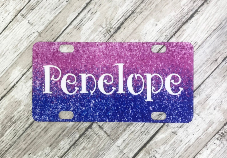 K&ouml;nnte beinhalten: Ein pinkfarbenes und blaues Glitzer-Kennzeichen mit dem Namen "Penelope" in wei&szlig;en Buchstaben.
