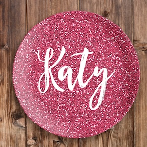 May include: A pink glitter round plate with the name "Katy" written in white cursive lettering.