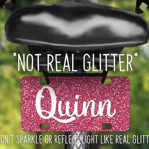May include: Pink glitter license plate with the name "Quinn" in white lettering. The text "*NOT REAL GLITTER*" and "*WON'T SPARKLE OR REFLECT LIGHT LIKE REAL GLITTER*" is printed below the plate.