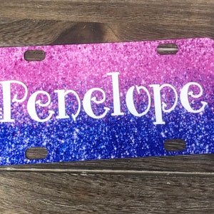 K&ouml;nnte beinhalten: Ein pinkfarbenes und blaues Glitzer-Kennzeichen mit dem Namen Penelope in wei&szlig;en Buchstaben.
