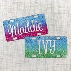 May include: Two personalised license plate covers with glitter backgrounds. The top cover is pink and blue with the name "Maddie" in white. The bottom cover is green and blue with the name "Ivy" in white.