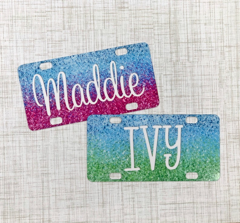 May include: Two personalised license plate covers with glitter backgrounds. The top cover is pink and blue with the name "Maddie" in white. The bottom cover is green and blue with the name "Ivy" in white.