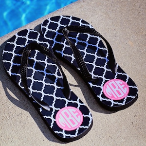 May include: A pair of black and white flip flops with a pink monogram on each. The monogram is a circle with the letters "MBE" inside.